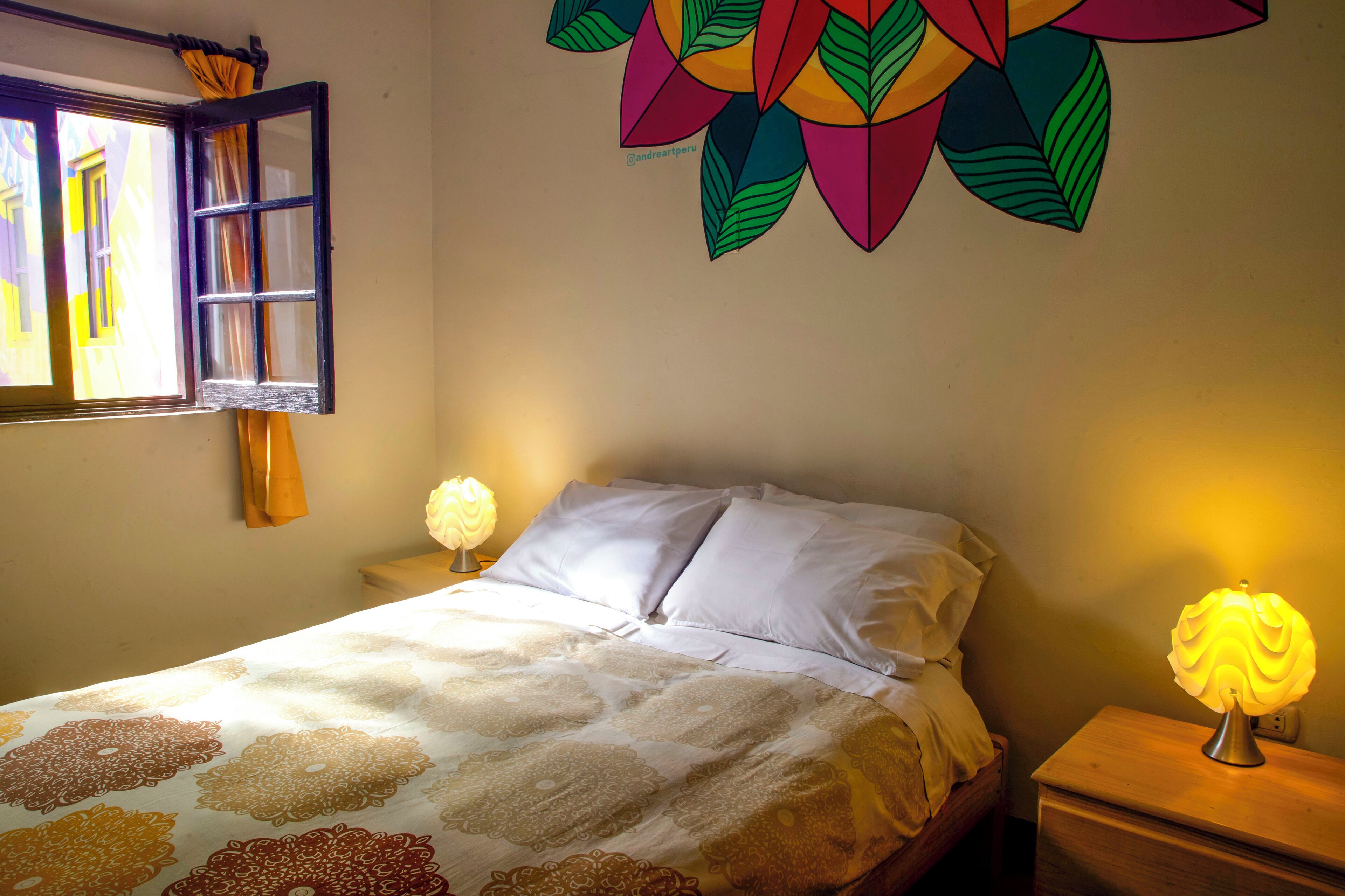 Ein Schlafzimmer mit Bett, Nachttisch, Wandlampe, einem Fenster mit Vorhängen und einer farbenfrohen Wanddekoration.