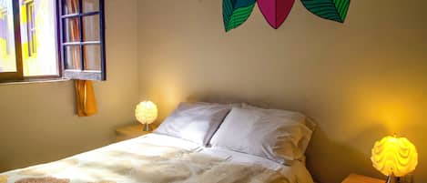 A bedroom with a bed, a nightstand, a wall-mounted lamp, a window with curtains, and a colorful wall decoration.