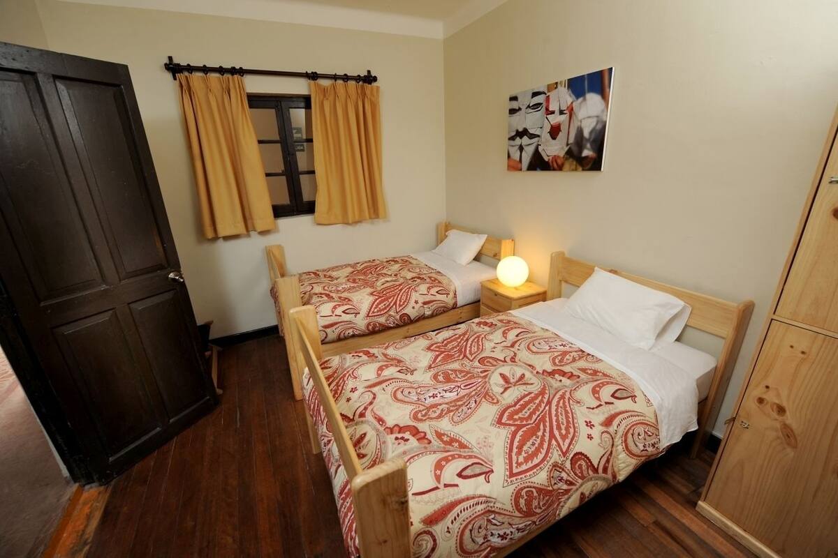A room with two beds, wooden headboards, a wardrobe, a window with curtains, and a wall-mounted picture.