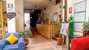 Un vestíbulo de hotel con un mostrador de recepción de madera, un sillón azul con un cojín amarillo, un sillón rojo y varias plantas en macetas.