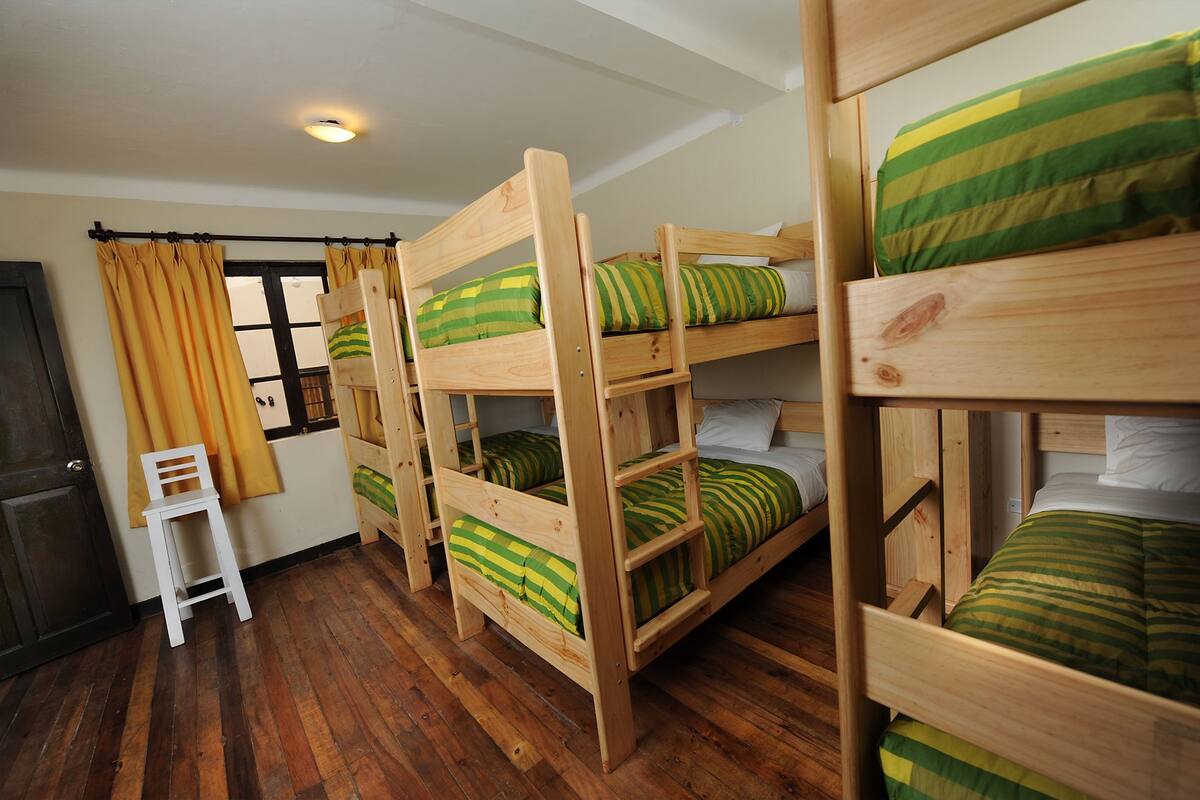 A room with bunk beds, wooden flooring, a small white chair, and a window with curtains.