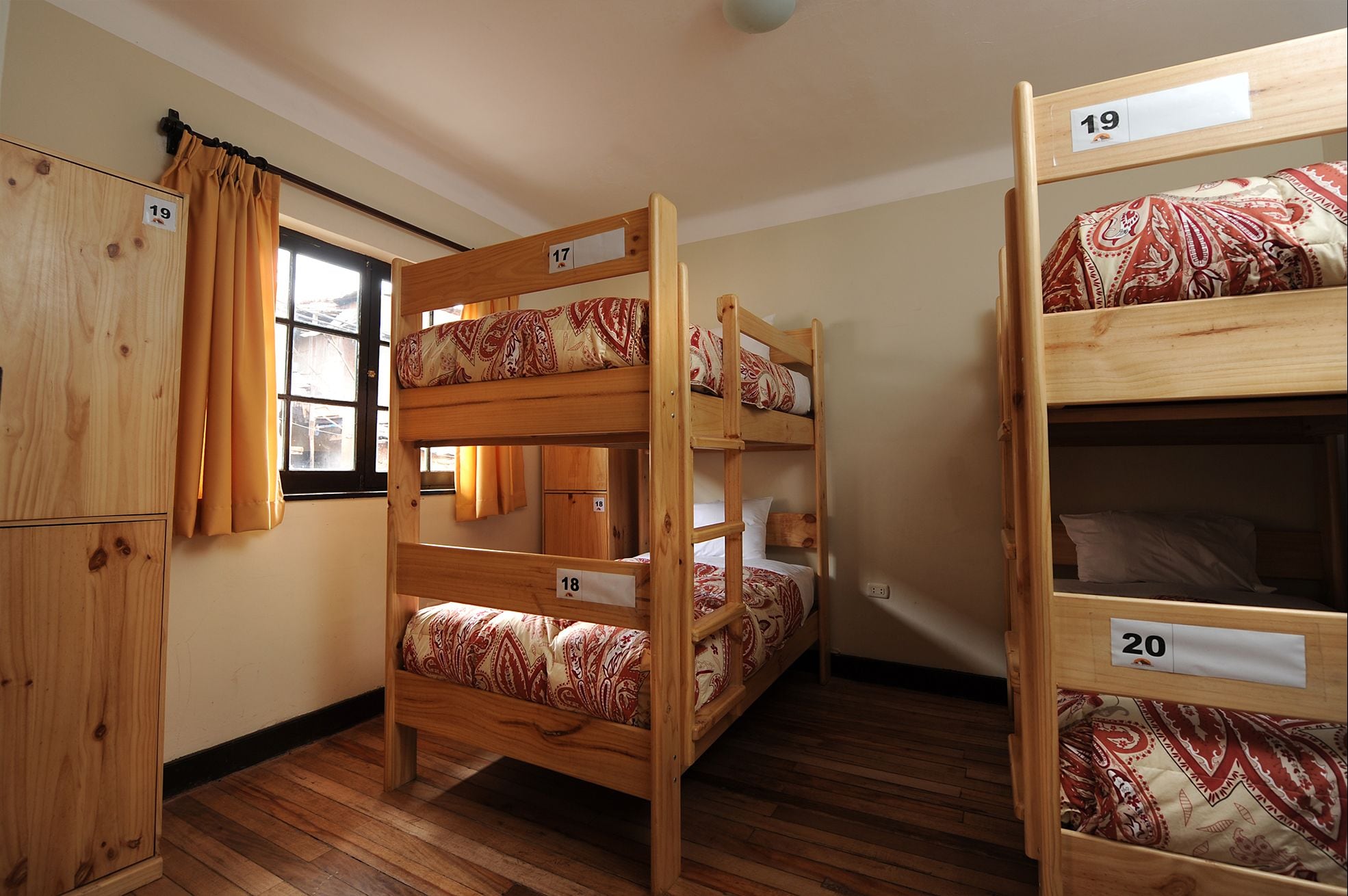 A dormitory room with bunk beds numbered 17 to 20, wooden furniture, and a window with curtains.
