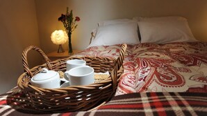 Una cama bien hecha con una colcha estampada, una bandeja de mimbre con tazas y una tetera, y un florero con flores sobre una mesita de noche.