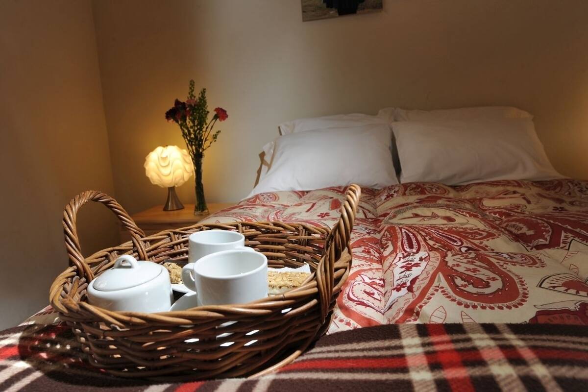 A neatly made bed with a patterned bedspread, a wicker tray with cups and a teapot, and a vase with flowers on a bedside table.
