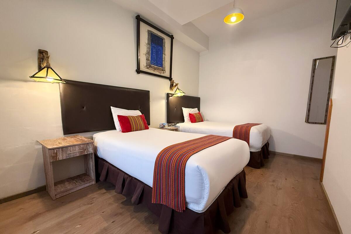 Ein Hotelzimmer mit zwei Betten, Holzboden, einem kleinen Nachttisch aus Holz, einem gerahmten Bild an der Wand und einer Deckenleuchte.