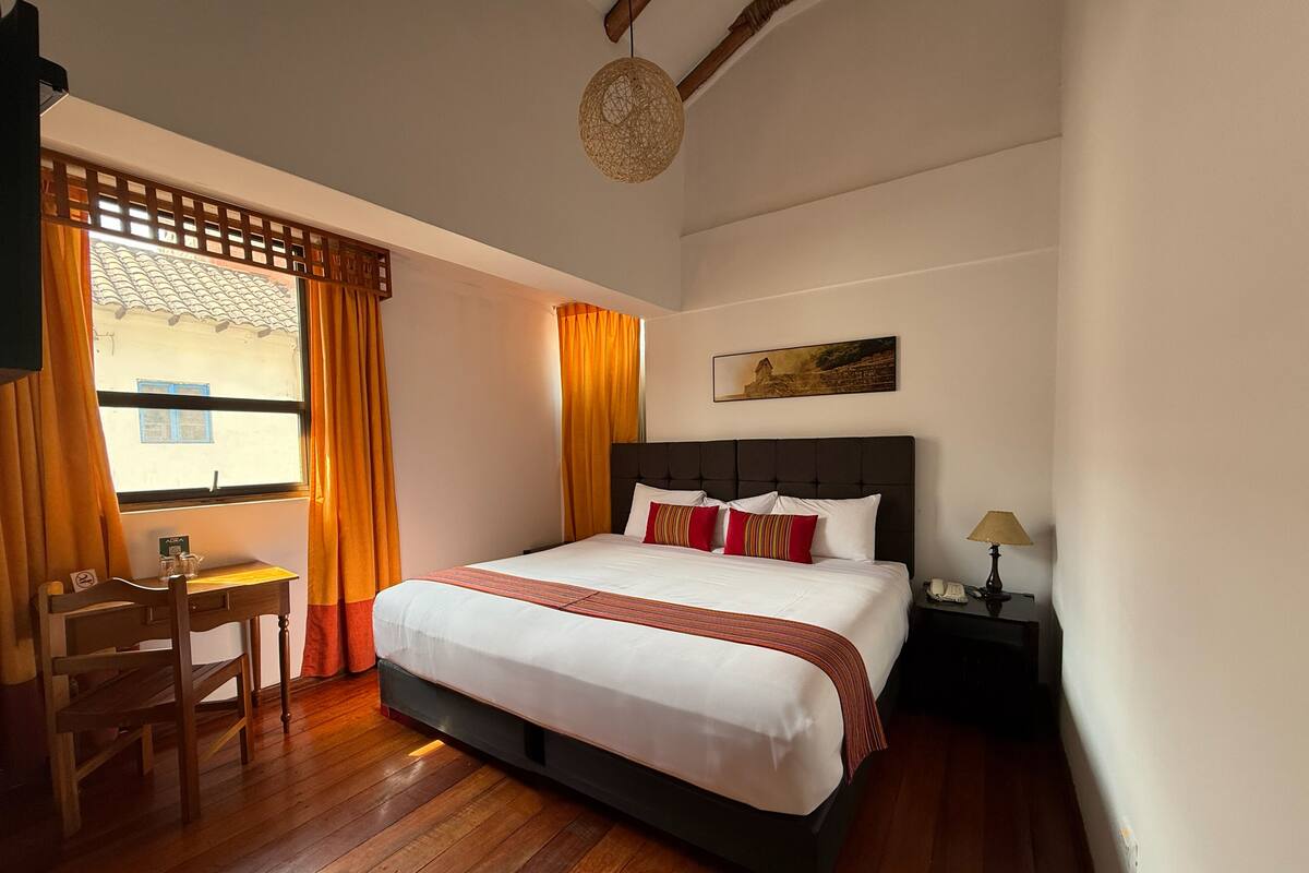Ein Hotelzimmer mit Bett, Schreibtisch, Stuhl, Nachttisch, Lampe und einem Fenster mit Vorhängen.