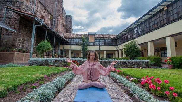 A person meditating on a blue mat in a courtyard with a stone building and a modern structure in the background. - Hotel Monasterio San Pedro (Cusco)