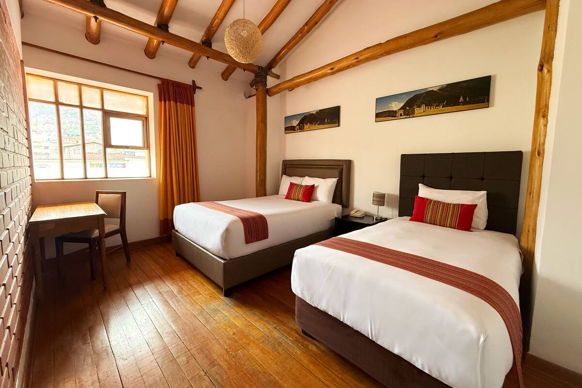 Ein Hotelzimmer mit zwei Betten, einem Schreibtisch, einem Fenster mit Vorhängen, Holzbalken an der Decke und Kunstwerken an den Wänden.