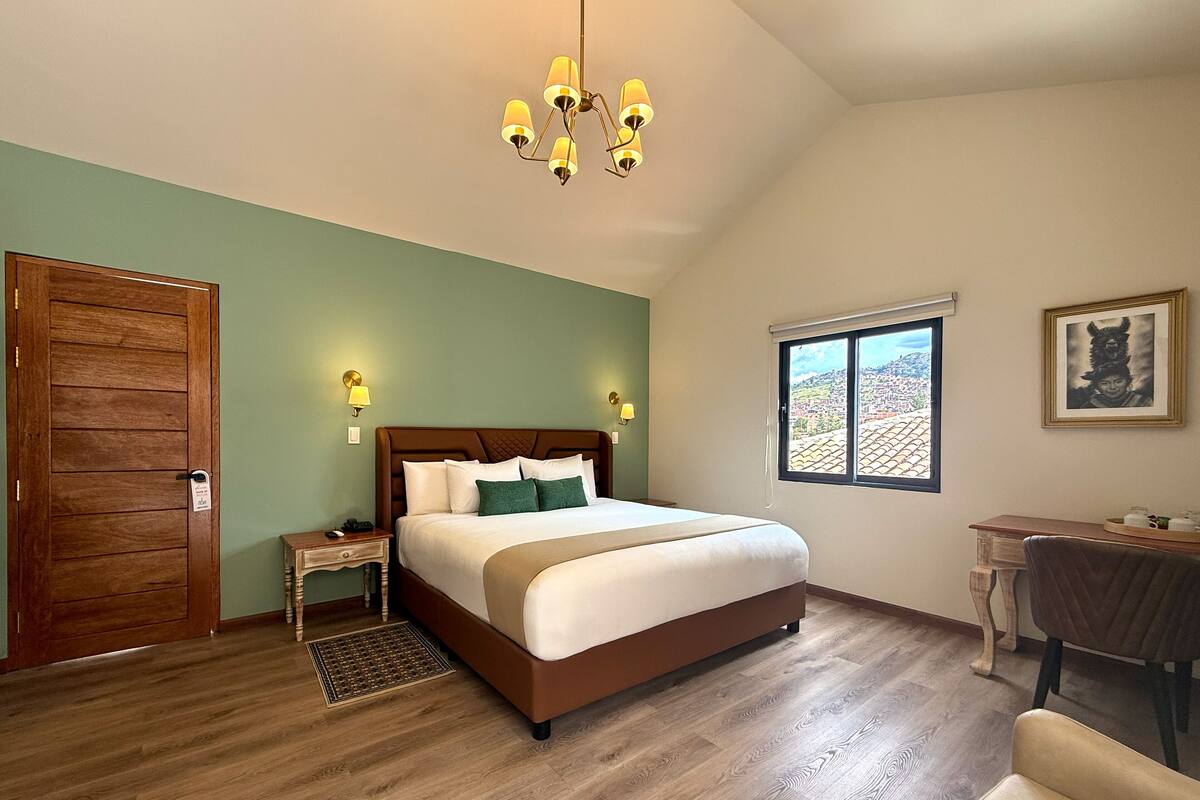 Ein Schlafzimmer mit Holztür, einem Bett mit weißer Bettwäsche, einem Schreibtisch und Blick auf eine Stadtlandschaft durch ein Fenster.