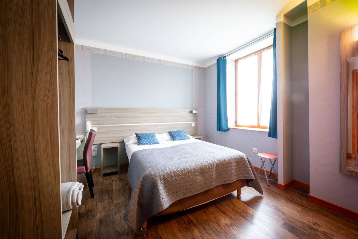 Ein Schlafzimmer mit einem hölzernen Bett, einem Sessel, einem kleinen Tisch und einem Fenster mit Vorhängen.