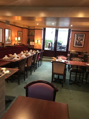 Un restaurante con una barra larga, varias mesas y sillas. Hay cuadros en las paredes y una puerta que da al exterior.