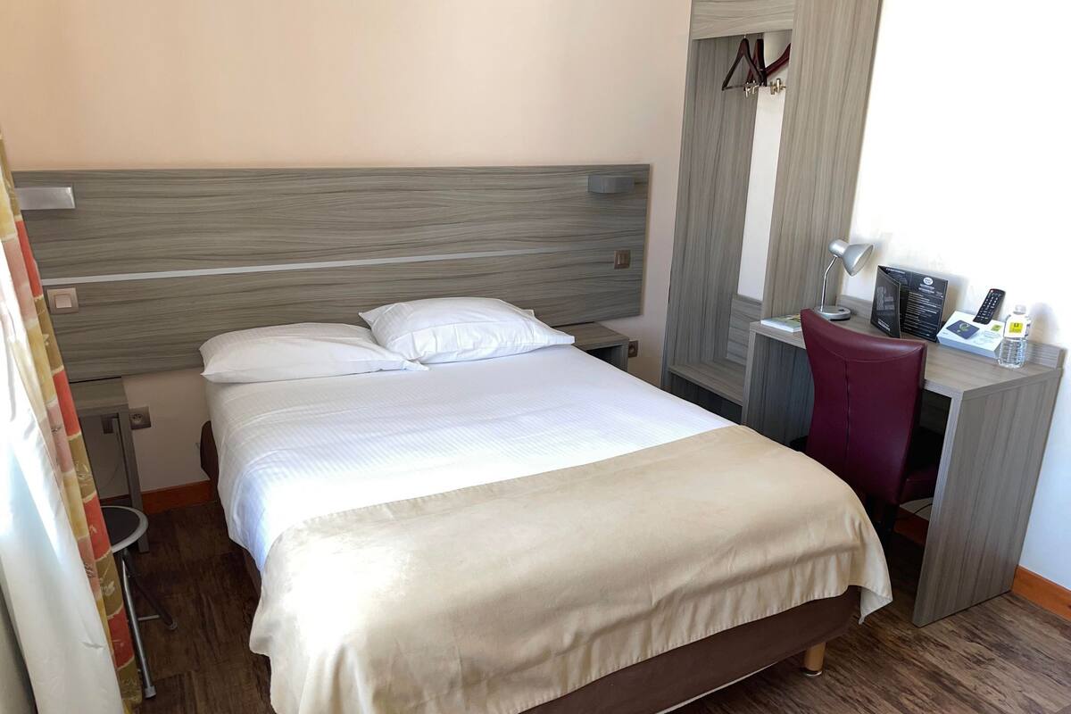 Ein ordentlich bezogenes Bett mit weißen Laken in einem Hotelzimmer, mit einem hölzernen Kopfteil und einem kleinen Schreibtisch mit Stuhl.