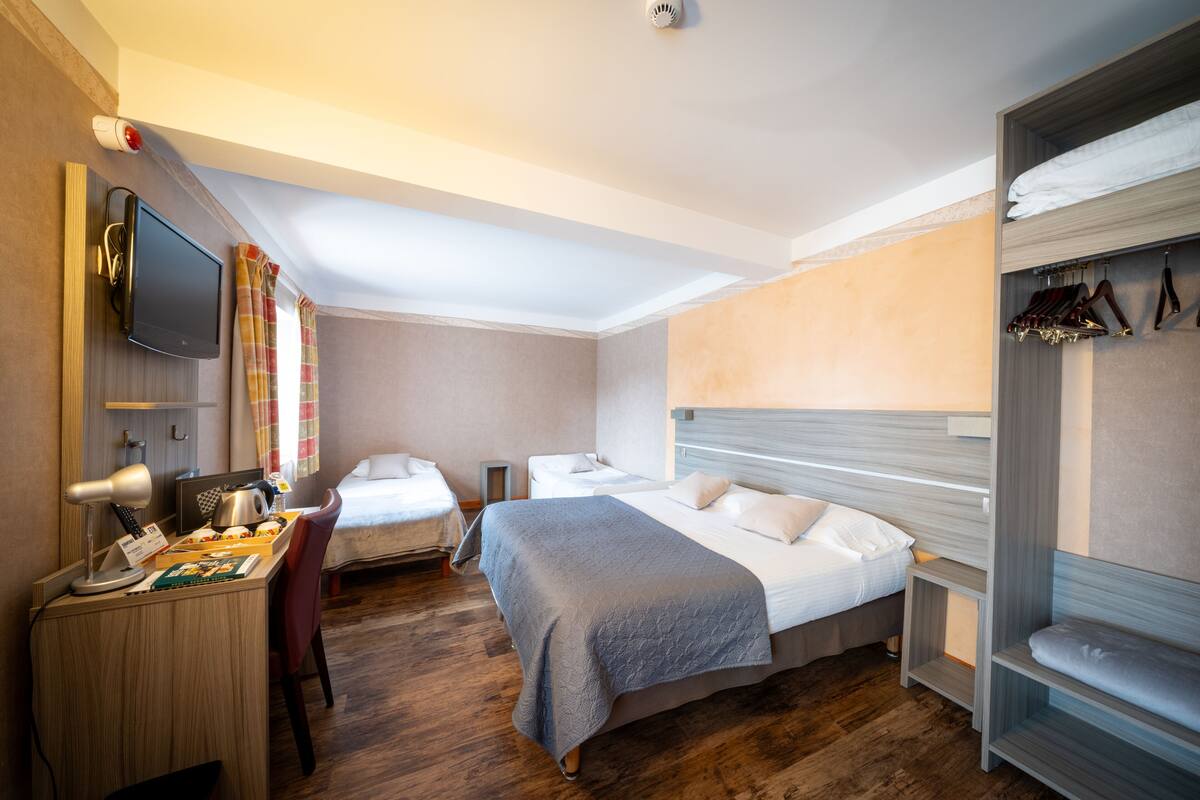Ein Hotelzimmer mit zwei Betten, einem Schreibtisch mit Computer, einem an der Wand montierten Fernseher und einem Kleiderschrank mit Kleidung.