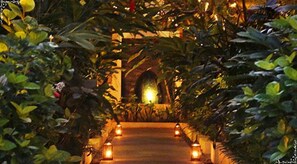 A pathway flanked by lush greenery and illuminated by lanterns, leading to a building with an arched entrance. - Arana Suite (Legian)