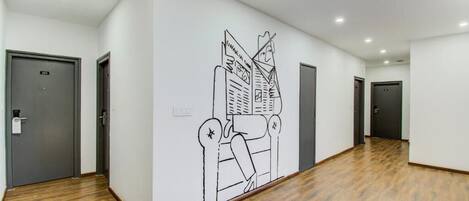 Un pasillo con un gran mural en la pared que representa un paisaje urbano, suelo de madera y puertas grises.