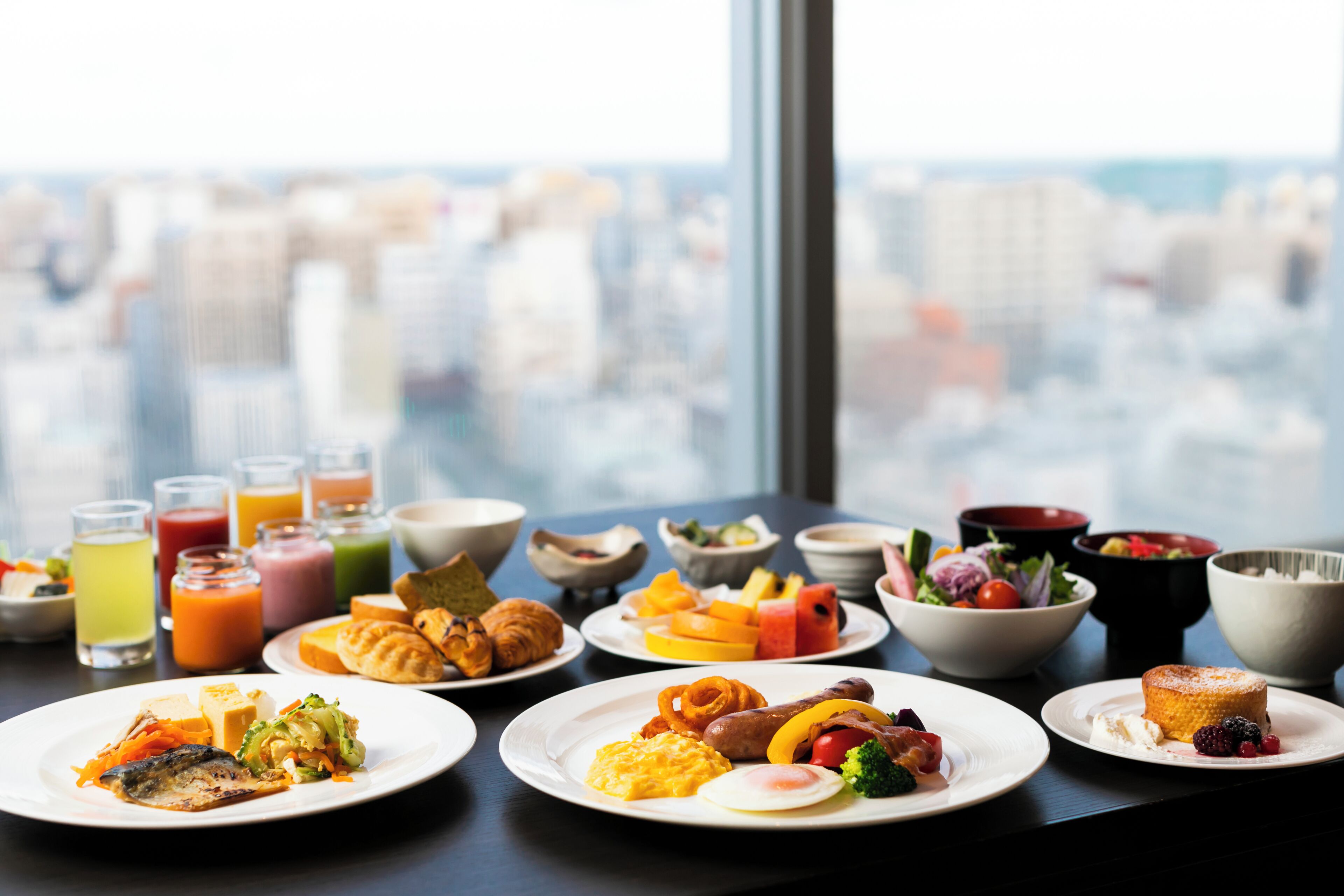 A breakfast buffet with various dishes and beverages on a table by a window overlooking a cityscape.