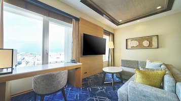 A hotel room with a large window, a desk, a flat-screen TV, a sofa, and a small round table.