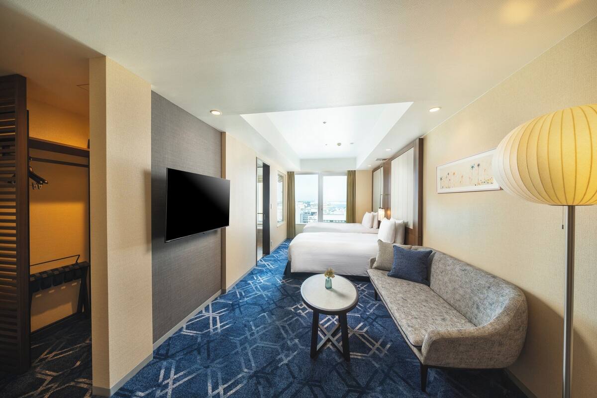 A hotel room with a bed, a sofa, a TV, a lamp, and a view of the city.