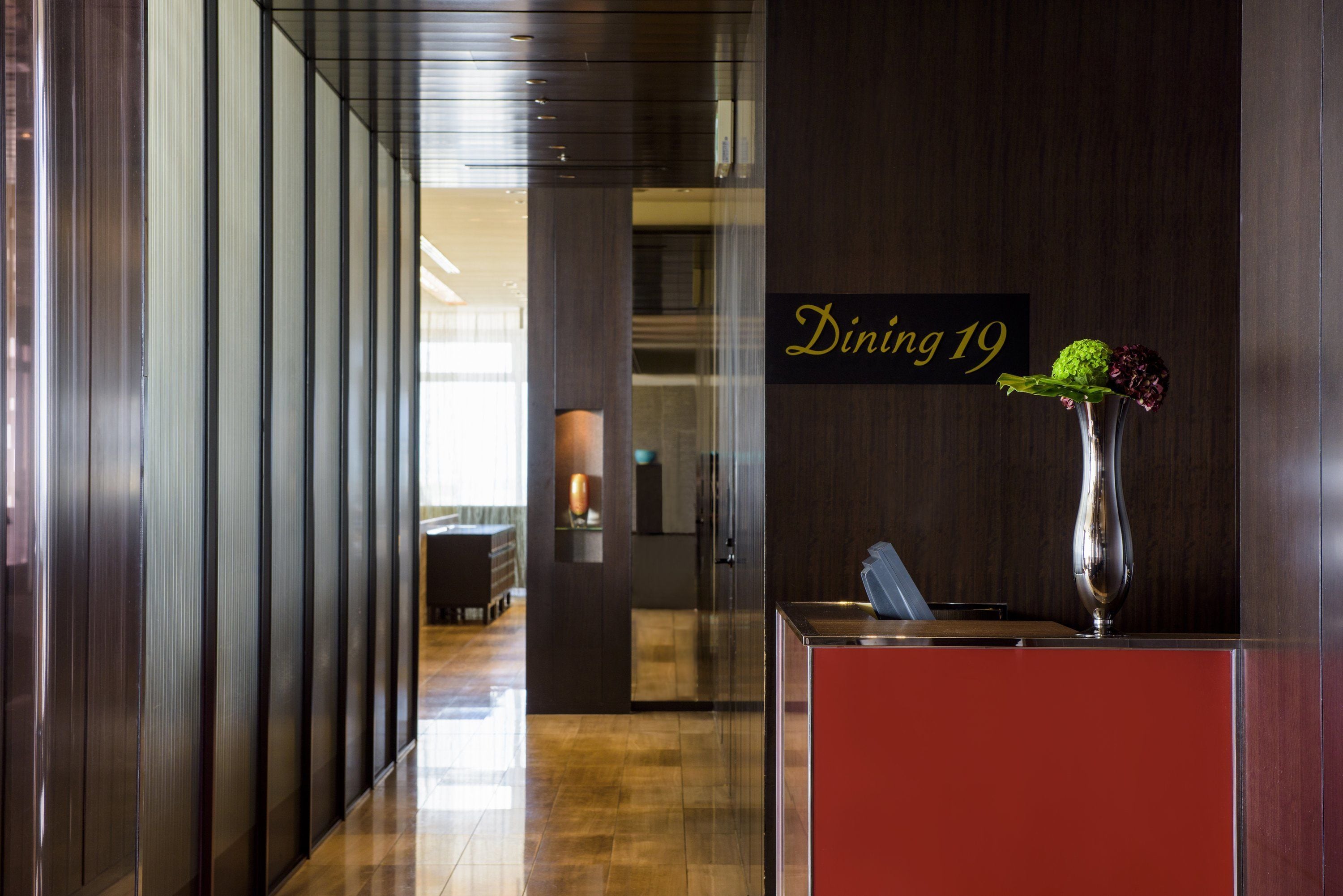 A modern hotel lobby with a reception desk, a sign indicating 'Dining 19', and a vase with flowers.