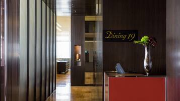 A modern hotel lobby with a reception desk, a sign indicating 'Dining 19', and a vase with flowers.