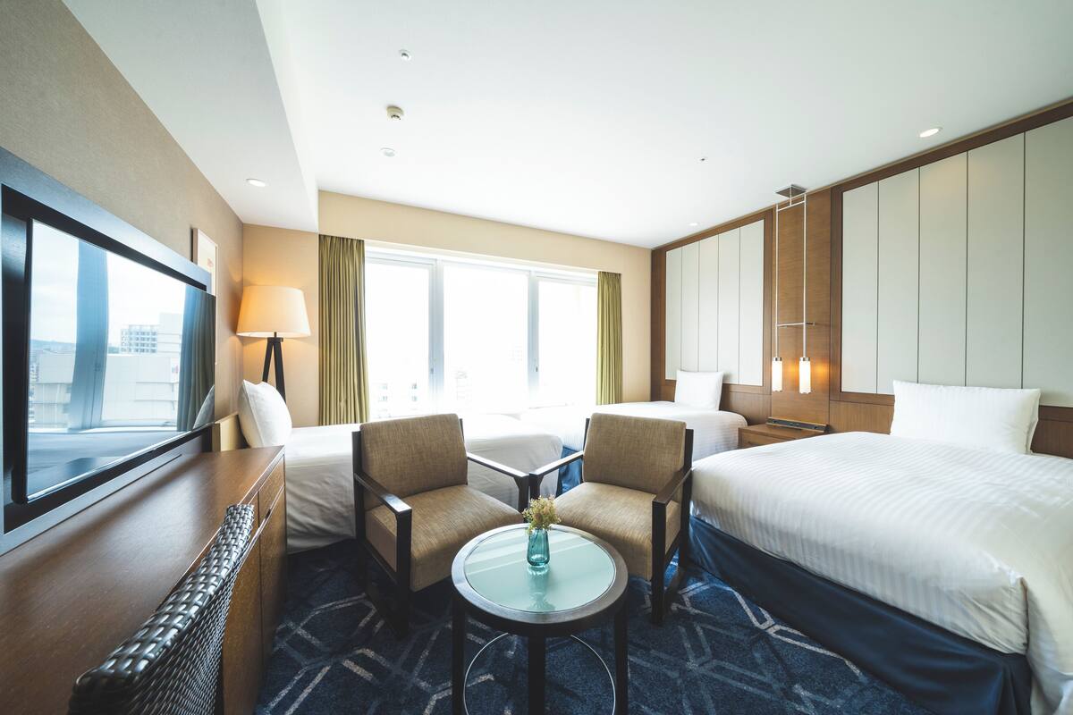 A hotel room with a large bed, two armchairs, a small table with a vase, and a large window with curtains.