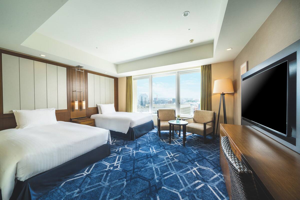 A hotel room with two beds, a TV, a desk, and a view of the city.