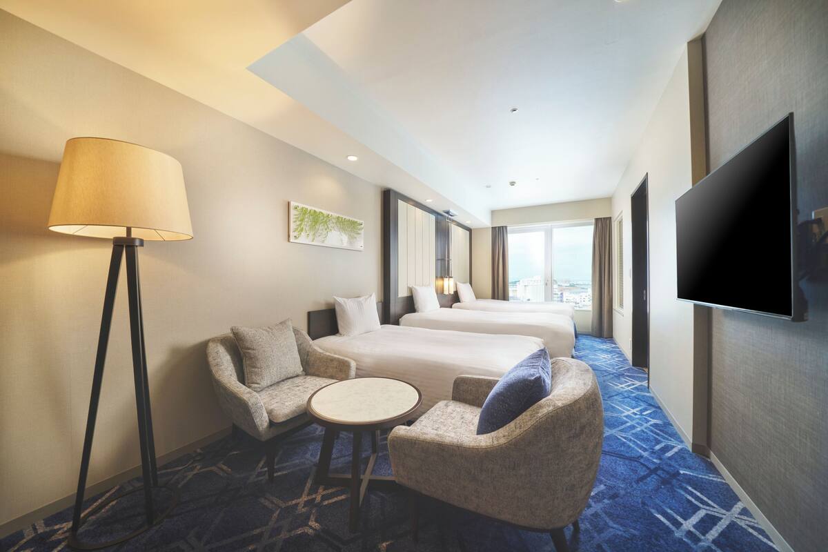A hotel room with a bed, two armchairs, a small table, a floor lamp, and a TV.