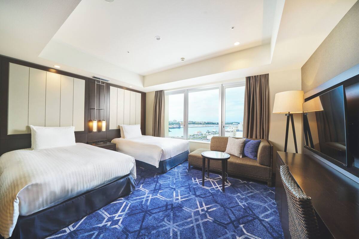 A hotel room with two beds, a sofa, a small table, a TV, and a large window with a city view.