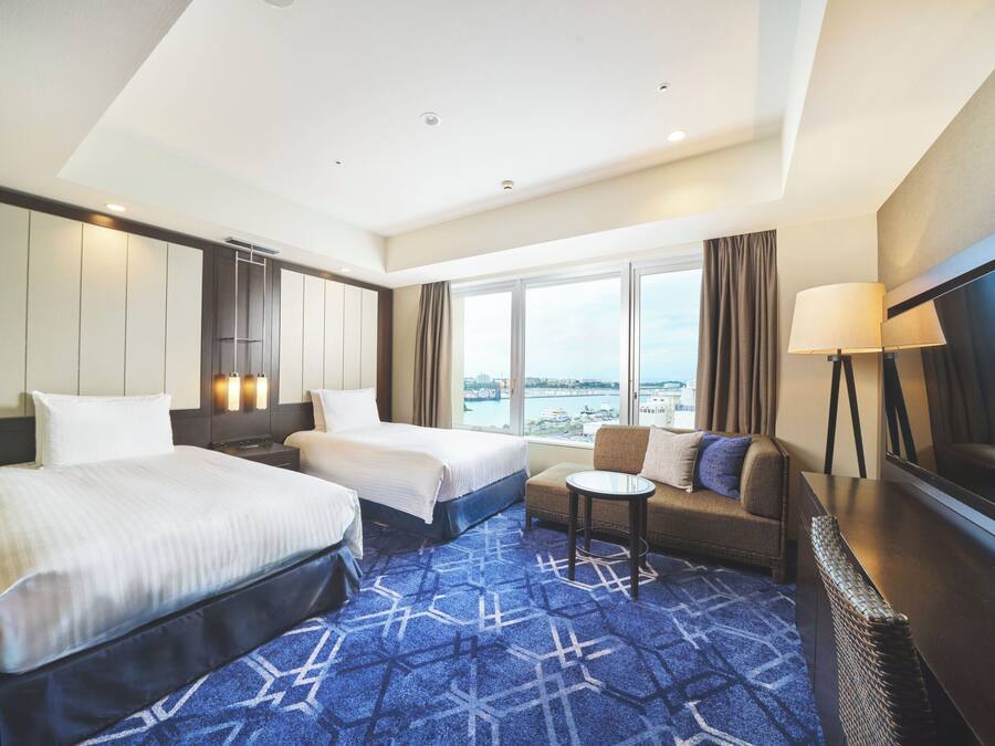A hotel room with two beds, a sofa, a small table, a TV, and a large window with a city view.