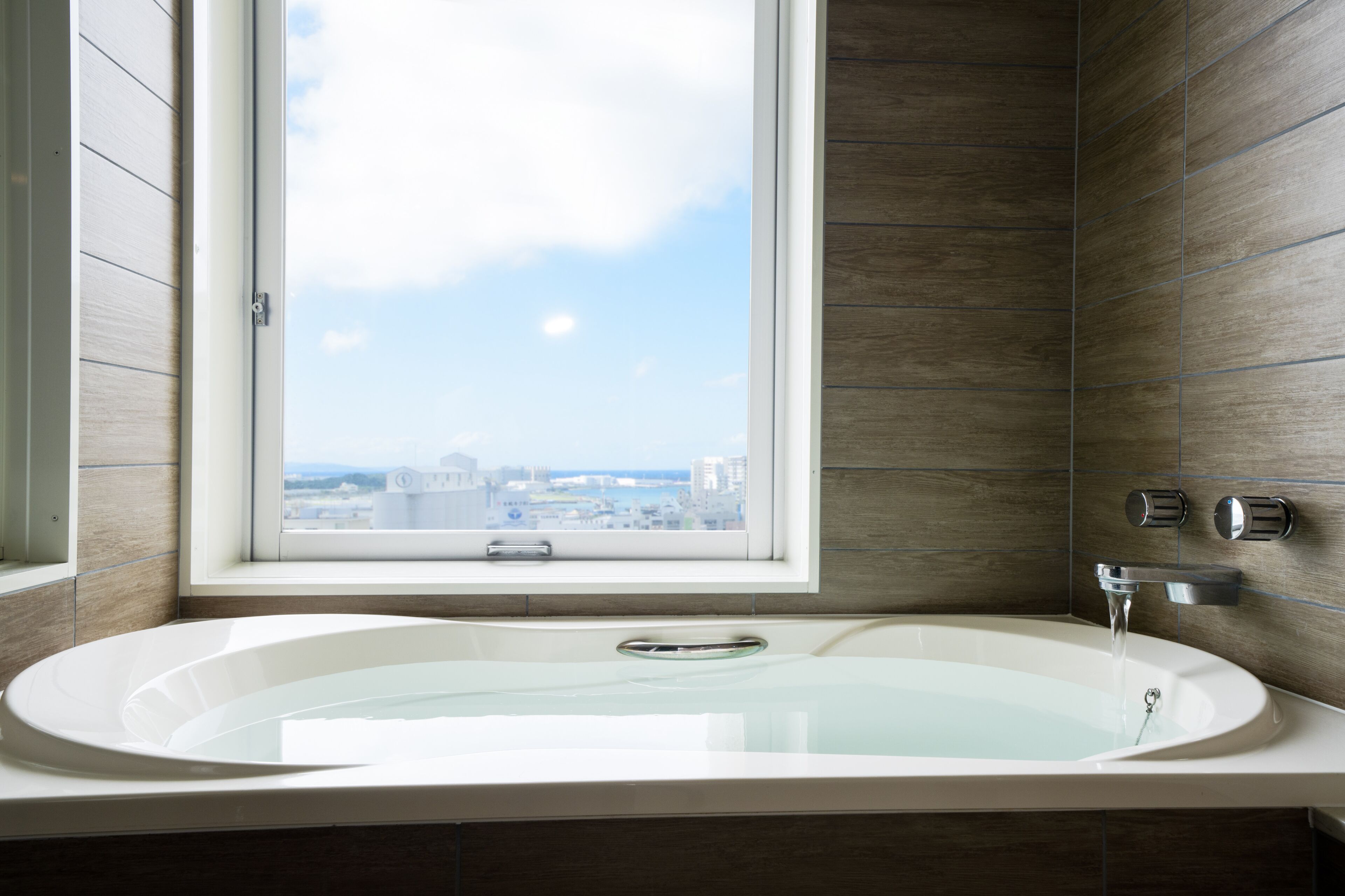 A modern bathroom with a large bathtub, a window with a city view, and wooden paneling.