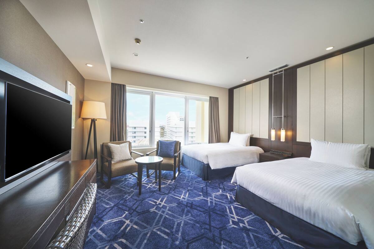 A hotel room with a large flat-screen TV, two beds, a sofa, a small table, and a view of the city through large windows.