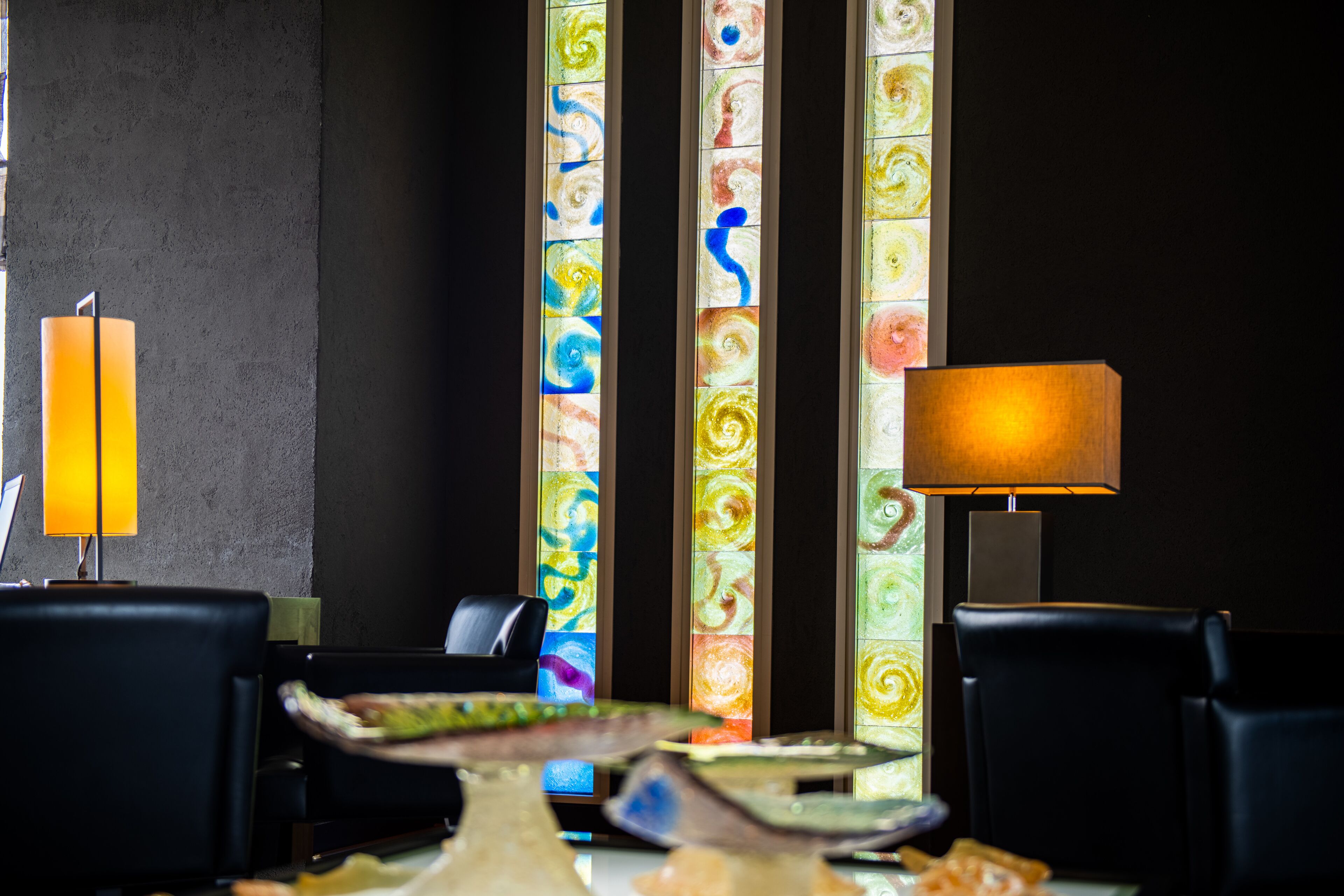 Un comedor con una mesa dispuesta para dos personas, una lámpara y un muro con paneles de vidrio emplomado de colores.