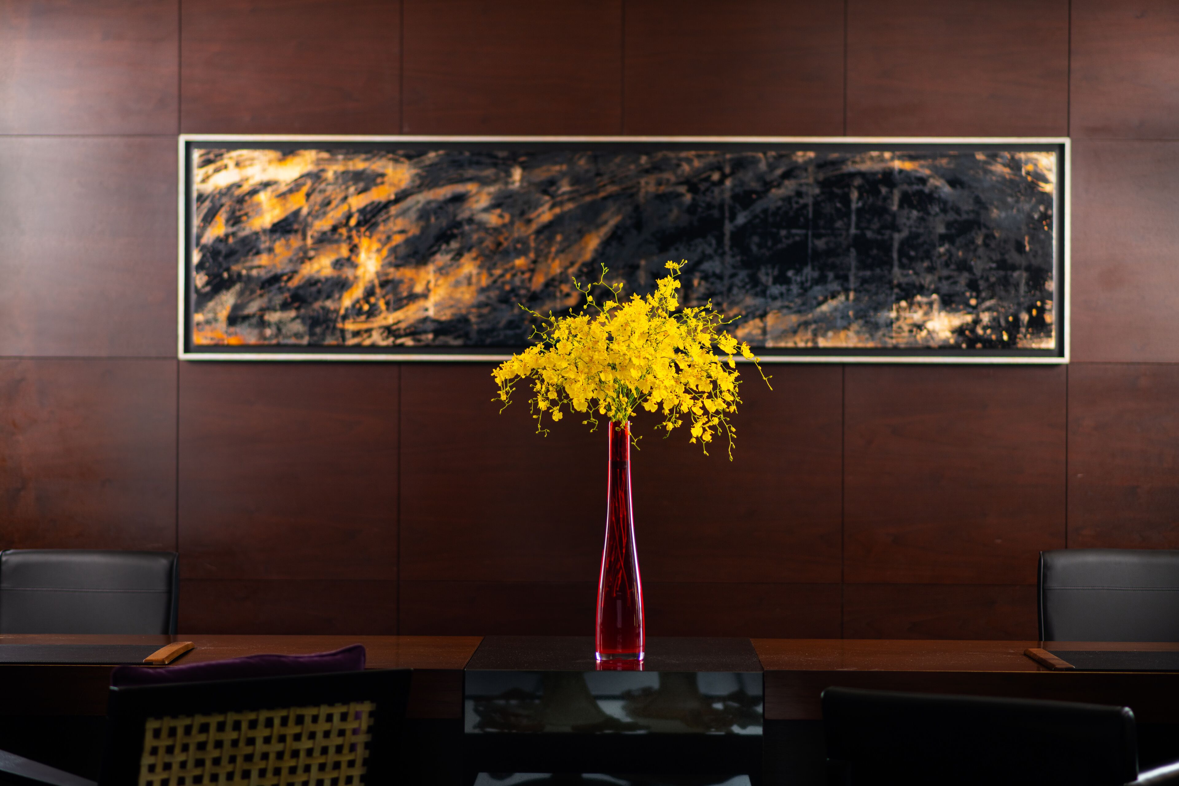 A room with a wooden panel wall, a framed abstract painting, a red vase with yellow flowers, and a black leather chair.