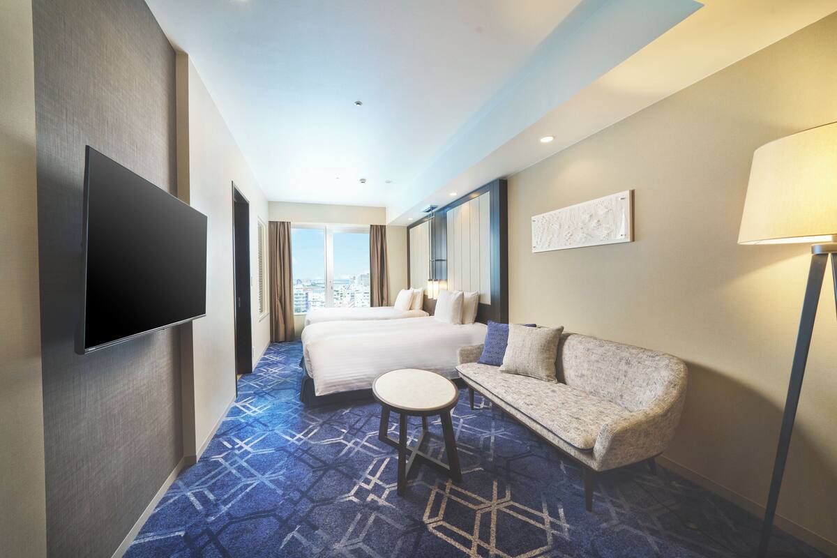 A hotel room with a large bed, a sofa, a small round table, a flat-screen TV mounted on the wall, and a city view through the window.