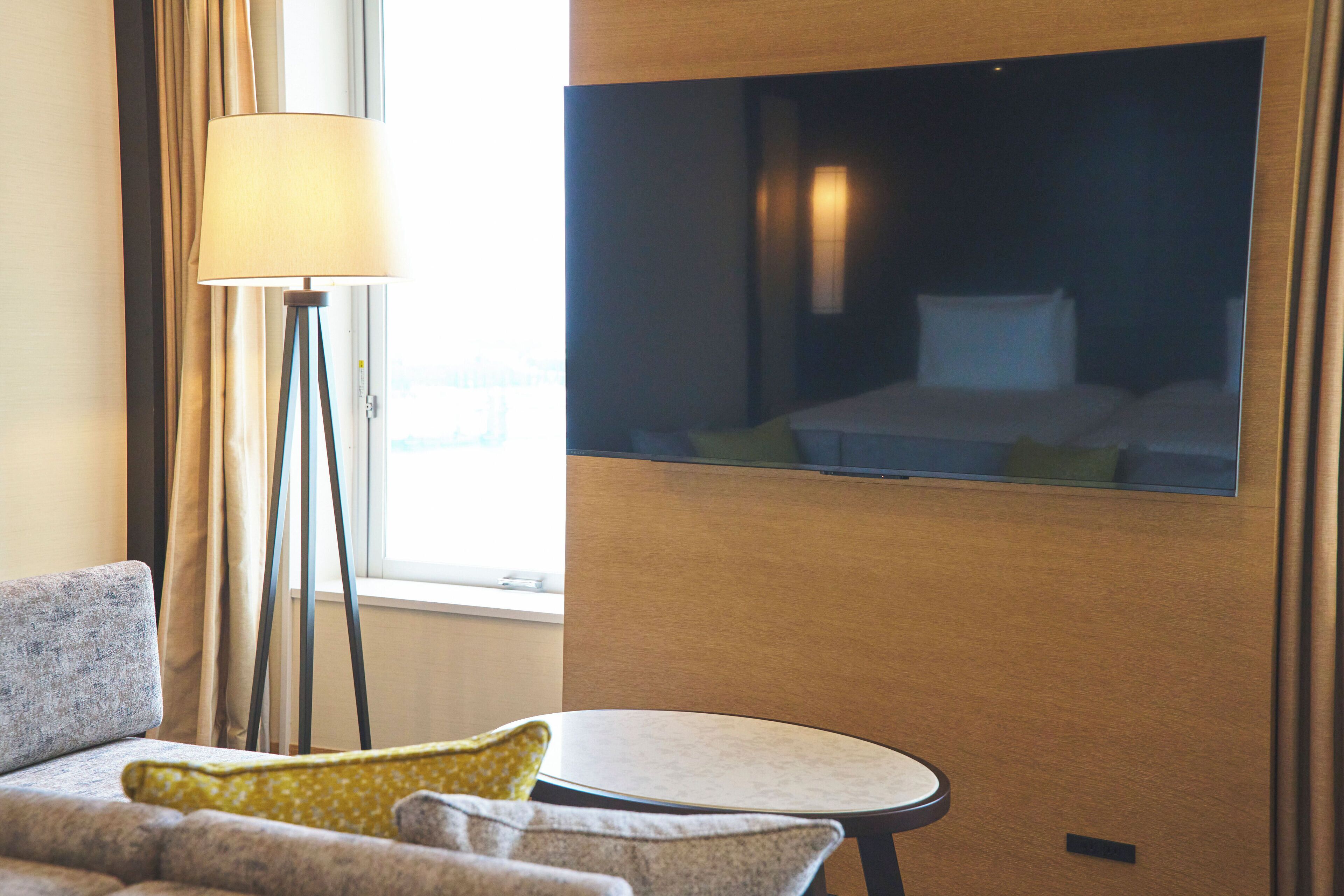 A hotel room with a sofa, a small round table, a floor lamp, and a TV mounted on the wall.
