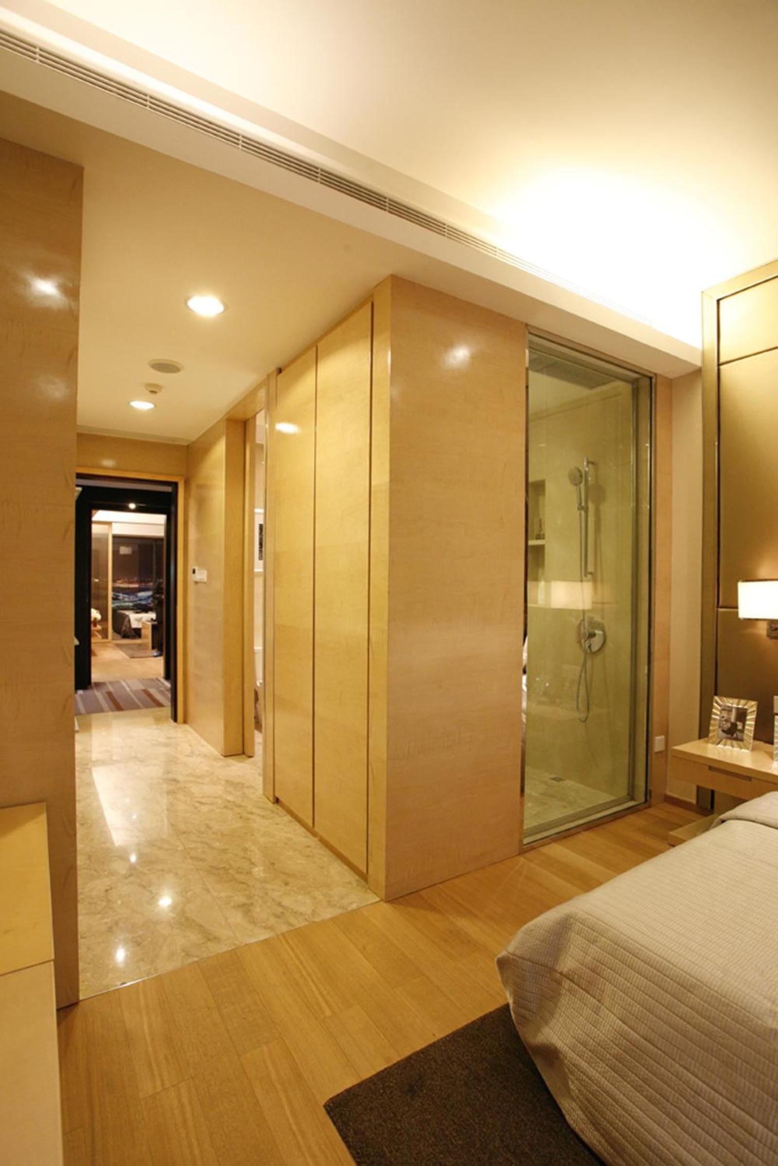 A hotel room with a bed, a glass-enclosed shower, wooden flooring, and a hallway leading to another room.