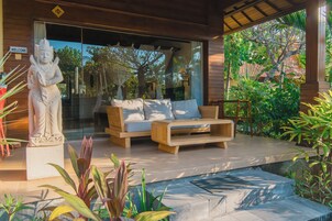 Un área de estar moderna al aire libre con un sofá y una mesa de madera, rodeada de exuberante vegetación y una estatua.