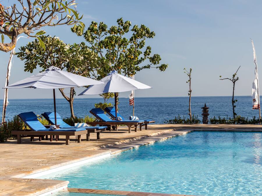 Zona de piscina con sillones reclinables y sombrillas, vista despejada del océano y un camino de piedra que conduce a la piscina.