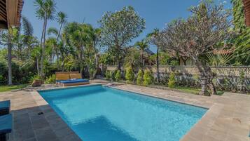 Una piscina con zona de descanso, rodeada de vegetación tropical y un muro de piedra.