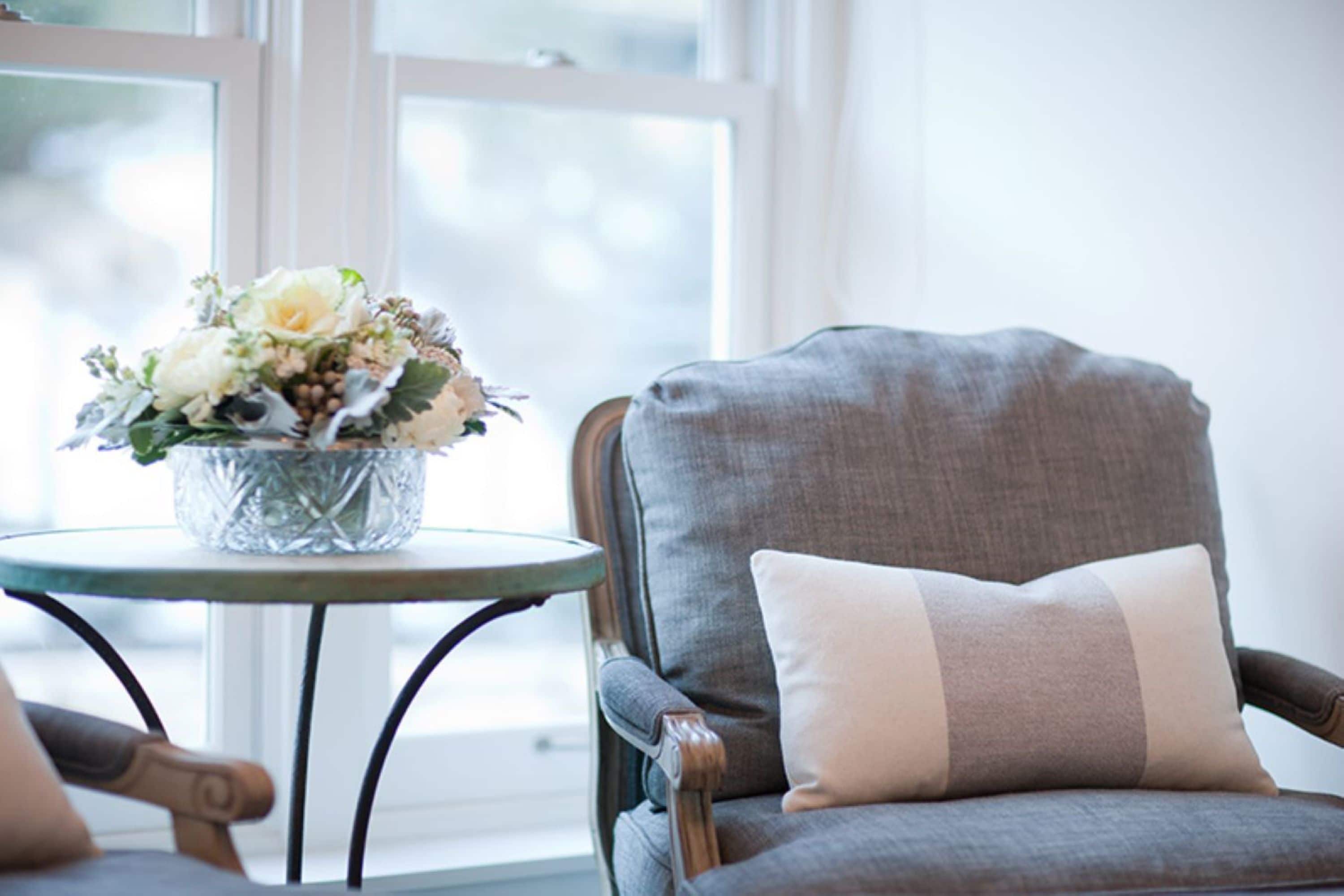 A chair with a cushion, a small round table with a vase of flowers, and a window with blinds.