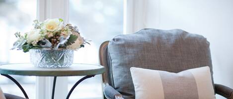 A chair with a cushion, a small round table with a vase of flowers, and a window with blinds.