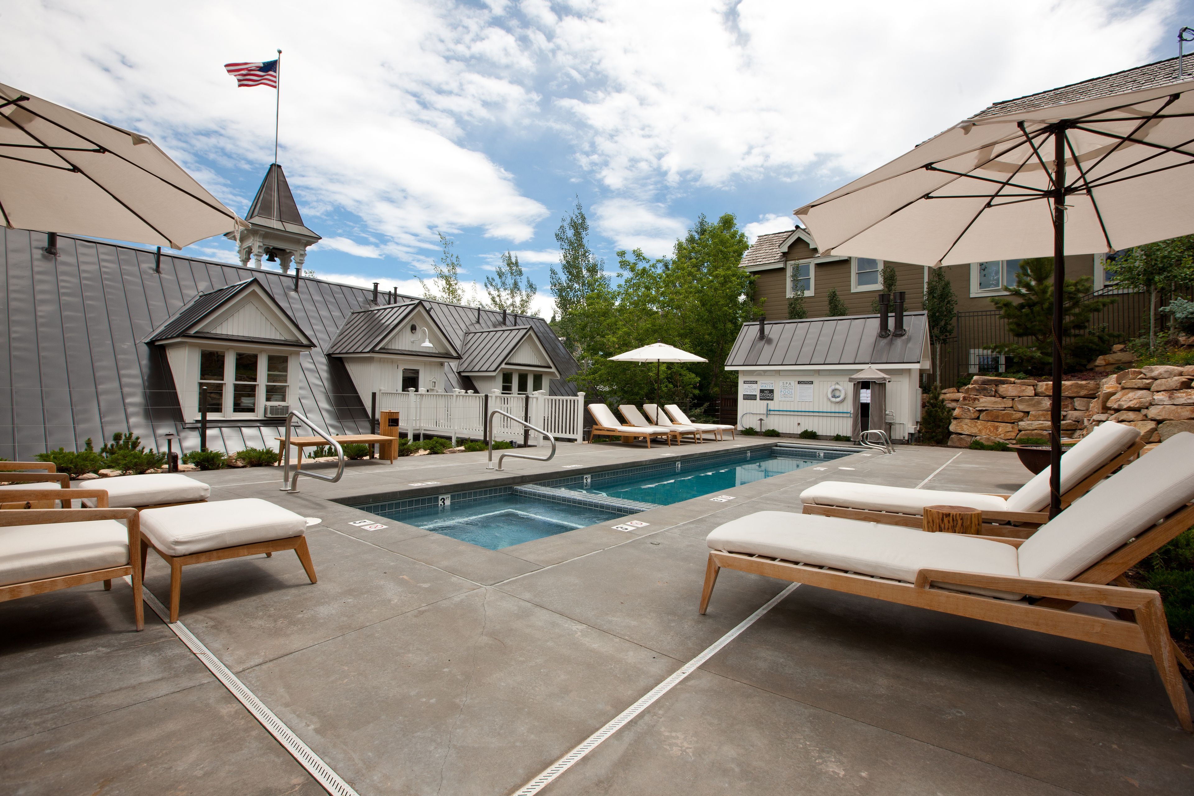 A pool area with lounge chairs and umbrellas, a flag, and a stone wall.