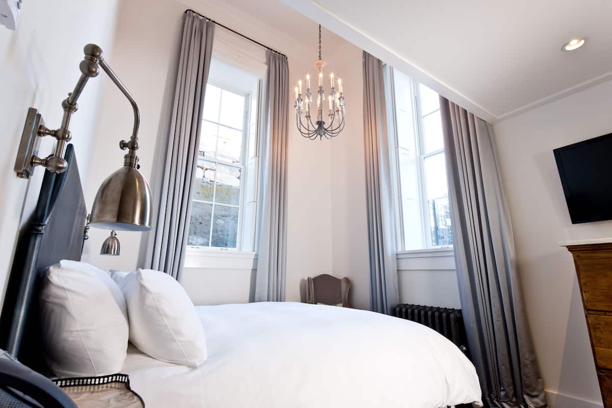 Habitación de hotel con una cama grande, un candelabro, un televisor y vistas de la ciudad a través de las ventanas.