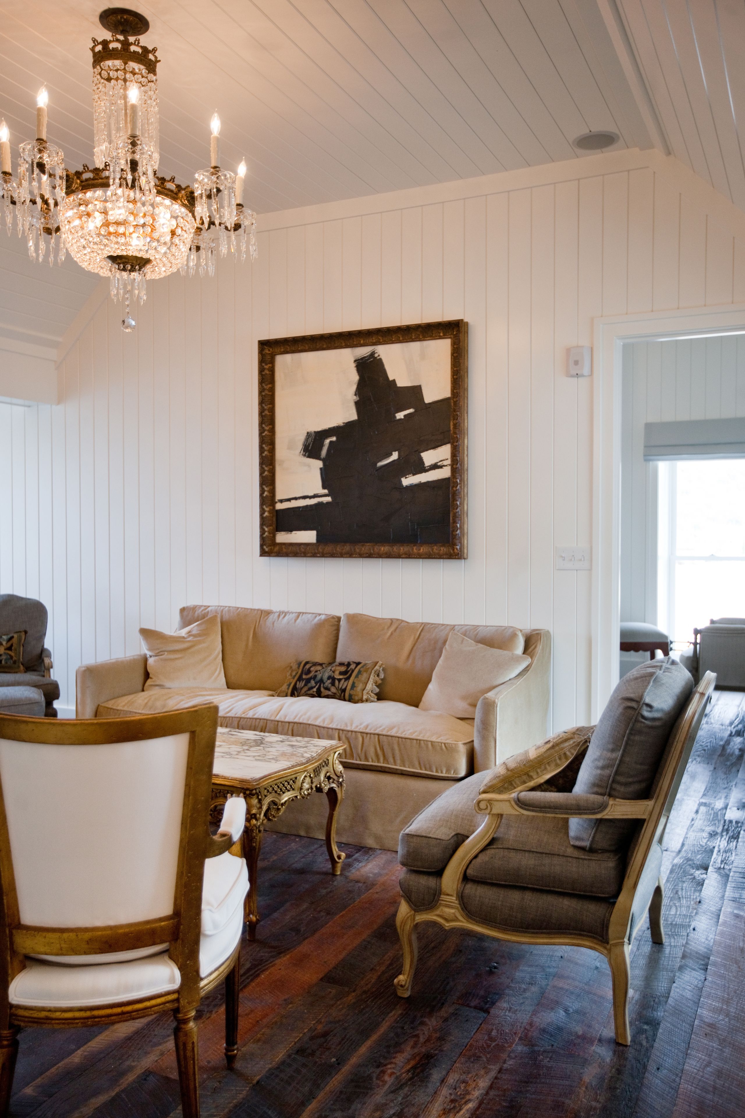 A room with a large chandelier, a framed abstract painting, a beige sofa, and a wooden floor.