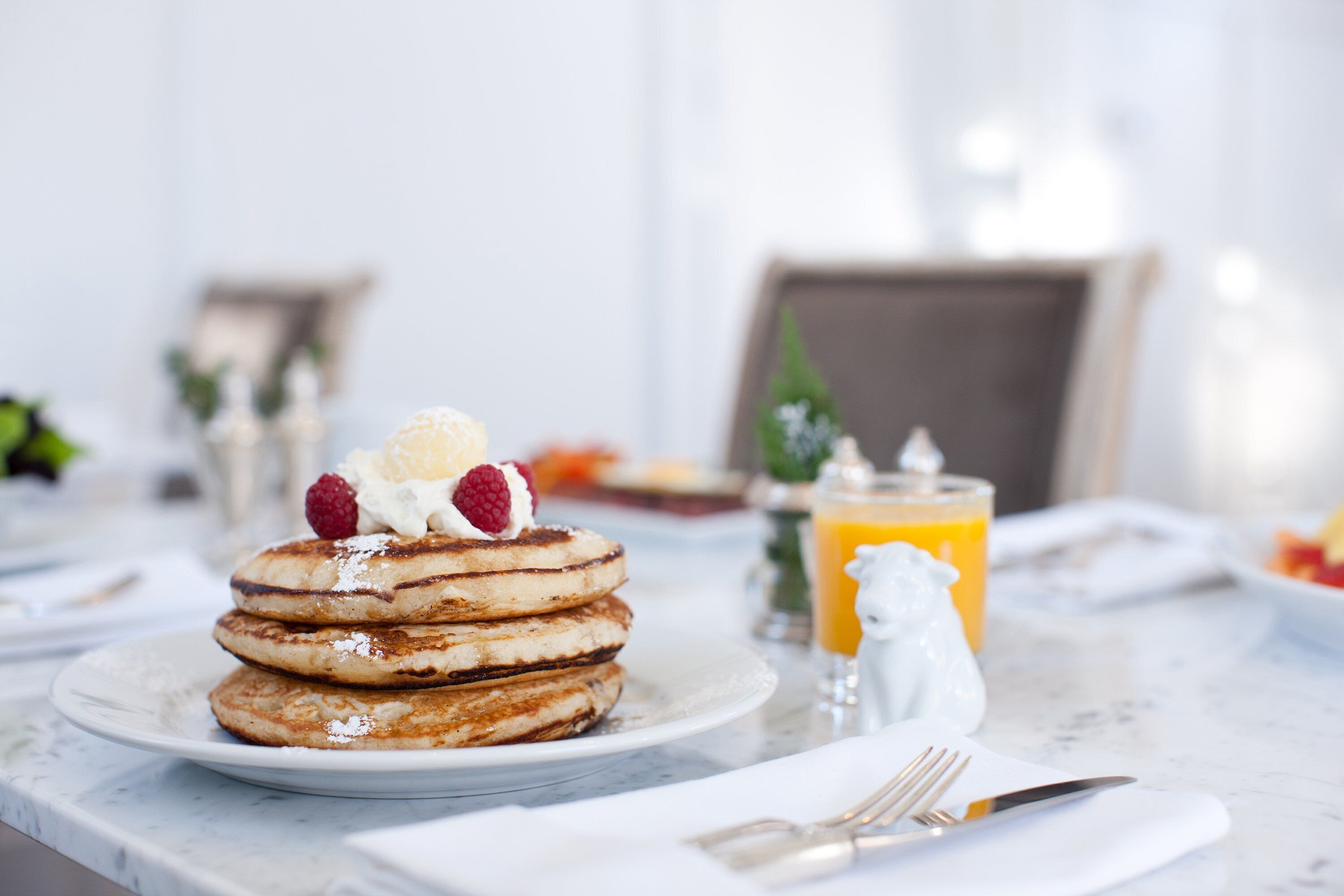 A breakfast setting with pancakes topped with whipped cream and berries, a glass of orange juice, and a salt shaker on a marble table.