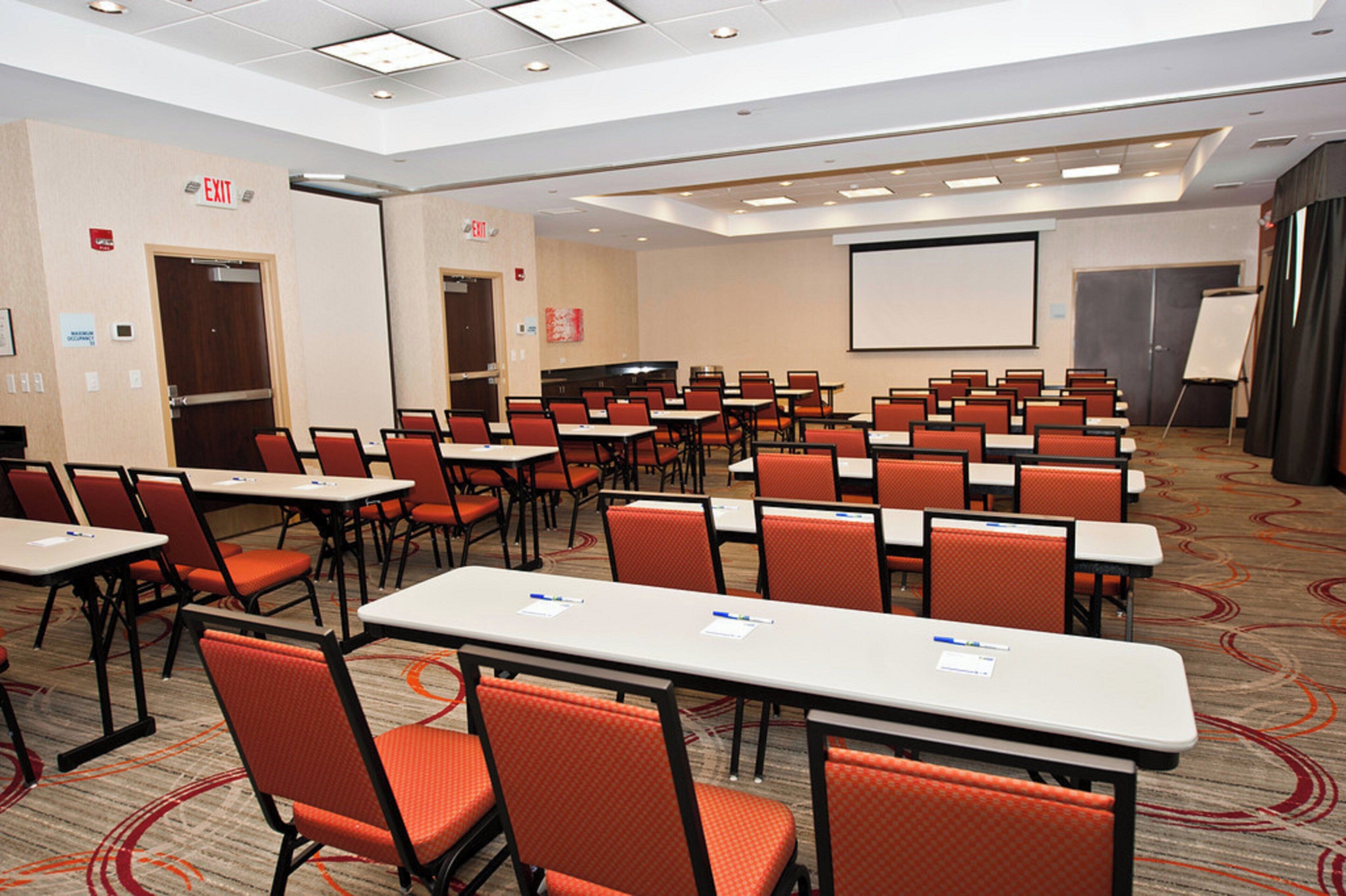 A conference room with rows of tables and chairs, a projector screen, and an exit sign.