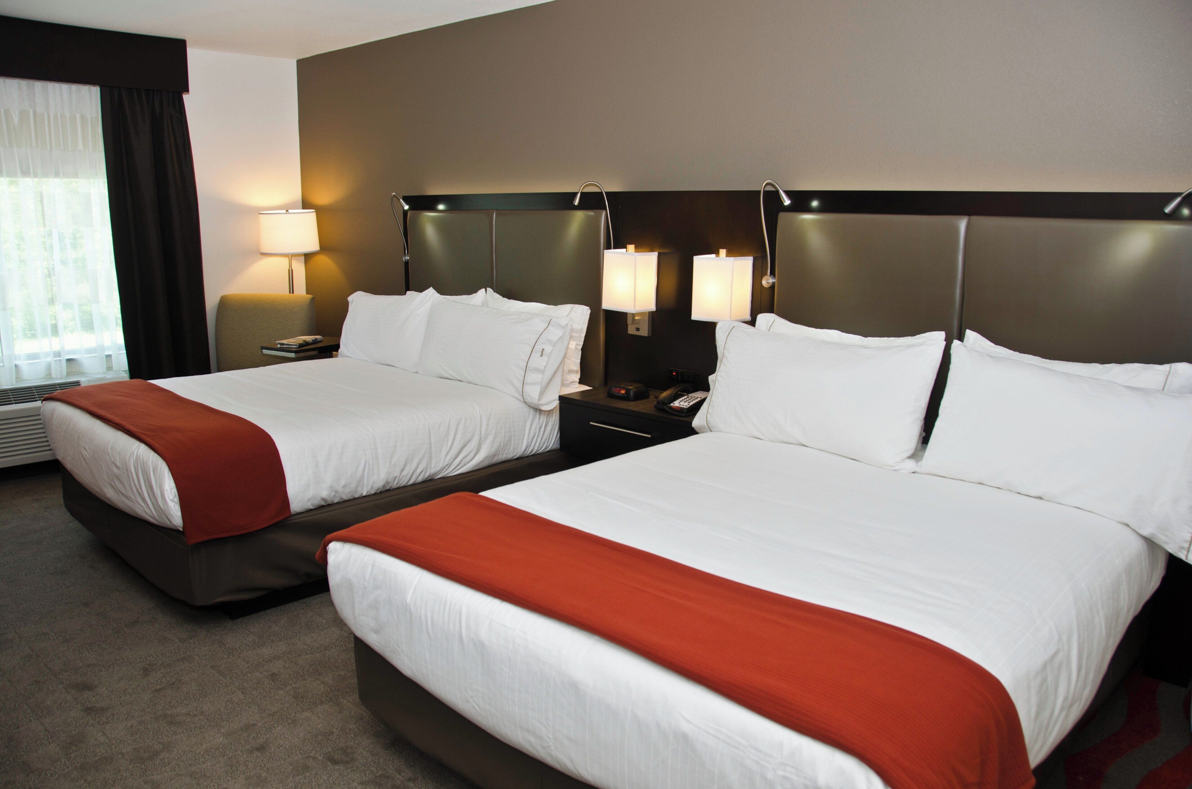 A hotel room with two beds, each with white bedding and red throw pillows. There are bedside tables with lamps and a remote control.