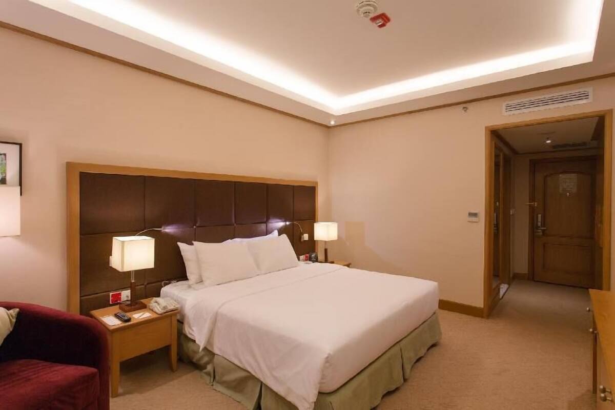 A hotel room with a large bed, two bedside lamps, a nightstand, a red armchair, and a door leading to another room.