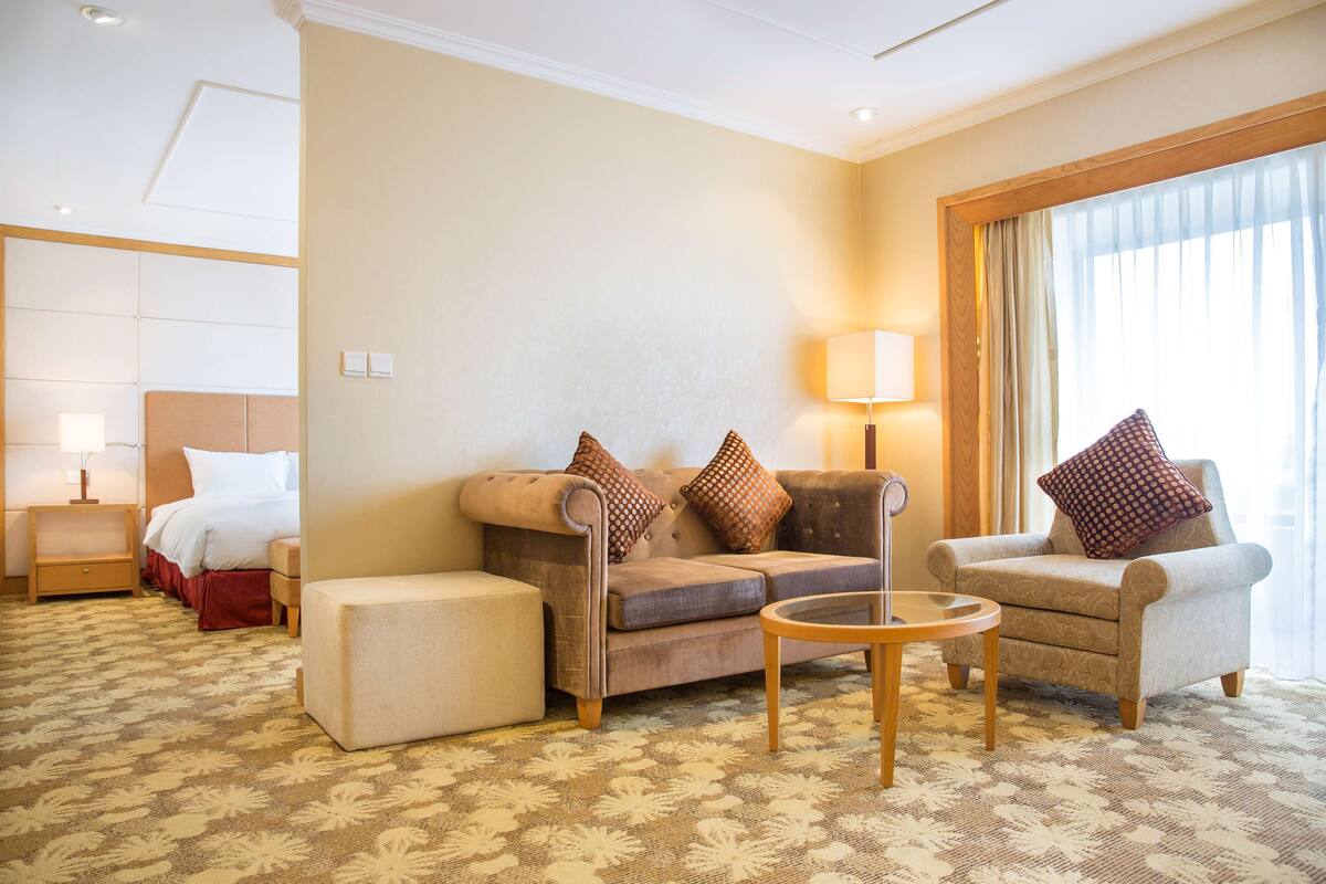 A hotel room with a sofa, armchair, ottoman, and a small table. There is a bed with a headboard and a nightstand with a lamp. The room has a carpeted floor and a large window with curtains.