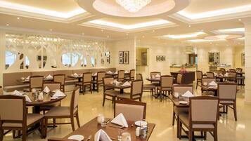 A spacious hotel dining area with multiple tables and chairs, a chandelier, and a decorative wall with tree-like patterns.